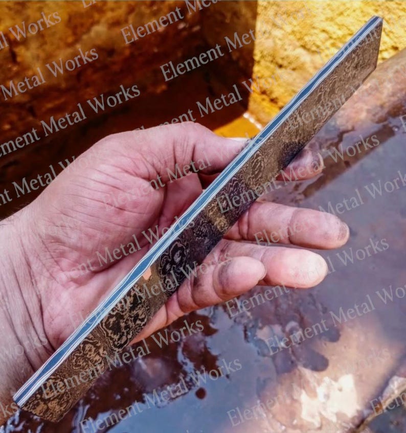 May include: A rectangular metal bar featuring a dark, intricate, etched floral pattern across its surface. The object displays a layered construction visible along its thin side, showcasing alternating light and dark metallic bands in a flat profile.