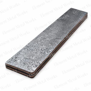May include: A rectangular metal bar featuring a dark, mottled, and textured gray surface with visible layers of copper and dark metal along the side edges. The surface has a distressed, weathered appearance with varying shades of charcoal and silver.