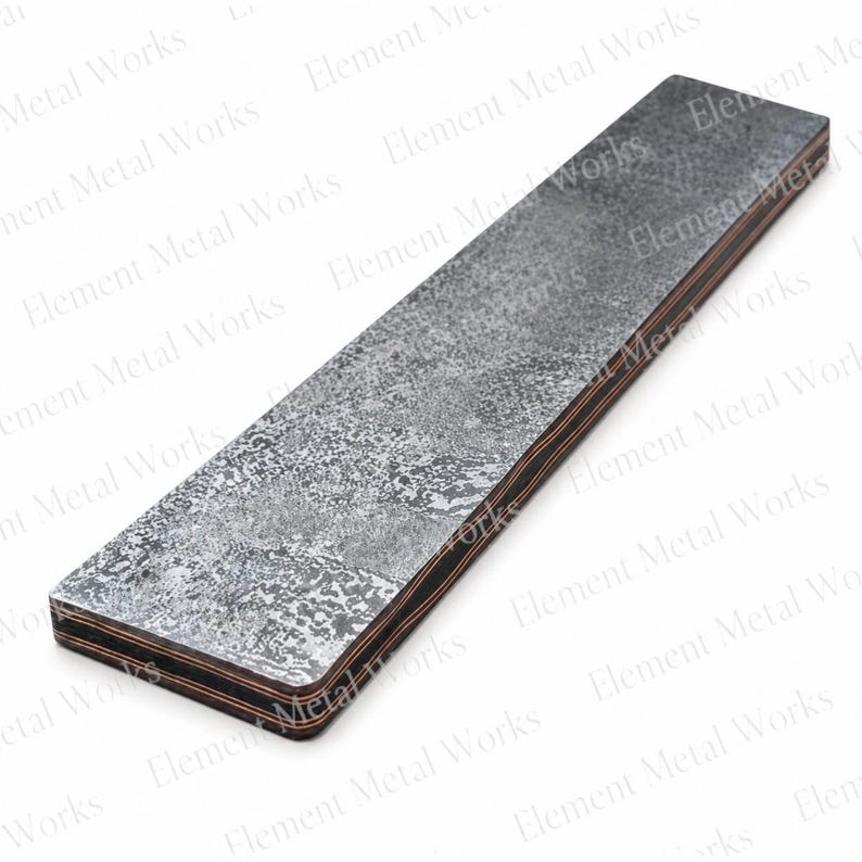 May include: A rectangular metal bar featuring a dark, mottled, and textured gray surface with visible layers of copper and dark metal along the side edges. The surface has a distressed, weathered appearance with varying shades of charcoal and silver.