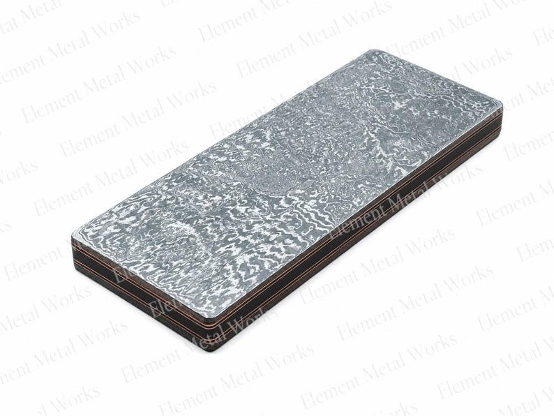 May include: A rectangular metal billet featuring a complex, swirling gray and silver Damascus pattern on the top surface, with visible layered construction along the sides showing alternating dark and copper-colored metallic bands.