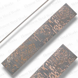 May include: A rectangular metal billet featuring a dark gray and copper-colored layered pattern known as mokume gane. The surface displays a wavy, wood-grain aesthetic created by the contrasting metal layers. A thin metal rod is positioned diagonally.