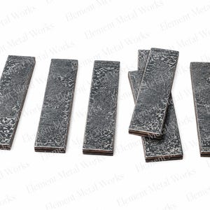 May include: Six rectangular metal bars with a dark, textured, hammered finish. The bars are arranged in a row, with one group of three overlapping slightly. Each piece has a distinct, rugged surface pattern and a layered appearance on the edges.