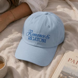 May include: A light blue baseball cap featuring the embroidered text Romance & RIGATONI in dark blue thread. The cap has a structured crown, a curved brim, and stitched eyelets on the top panels, providing a clean and simple design aesthetic.