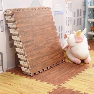 May include: Interlocking foam floor mats with a wood grain pattern in shades of brown and tan. The mats are stacked against a wall and also cover the floor. A plush unicorn toy with a pink mane and horn is in the background.