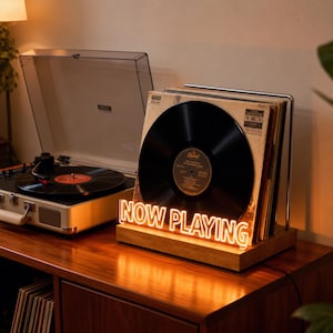 May include: A cream-coloured record player with a clear lid is next to a wooden stand holding vinyl records. A neon sign reading "NOW PLAYING" lights up the records. The setup is on a wooden cabinet.