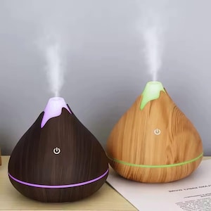 Op de afbeelding: Twee druppelvormige diffusers voor etherische olie met een houtnerf afwerking. De linker diffuser is donkerbruin met een paars licht aan de top en basis, terwijl de rechter diffuser lichtbruin is met een groen licht. Beide verspreiden nevel.