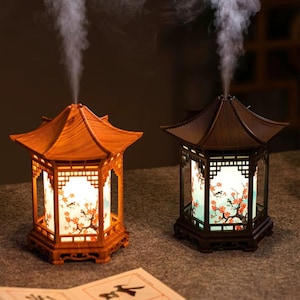 May include: Two wooden essential oil diffusers in a pagoda design. One is light brown, the other dark brown. Both have a floral design on the inside and emit a stream of vapor. The diffusers sit on a neutral surface.