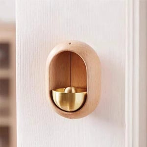 May include: A wooden wall-mounted bell with a brass-coloured bowl and a small ball-shaped clapper. The bell is encased in a light-coloured, oval-shaped wooden frame. The bell is mounted on a white wall.