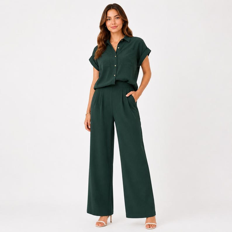 May include: A dark forest green jumpsuit featuring a button-down shirt top with short cuffed sleeves, a collared neckline, and a single chest pocket. The bottom consists of wide-leg trousers with front pleats and side pockets in a matching fabric.
