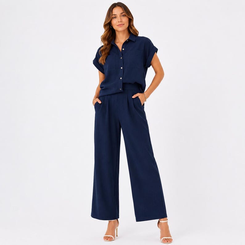 May include: A navy blue jumpsuit featuring a collared button-down shirt top with short cuffed sleeves and wide-leg trousers. The garment includes a front button placket, a chest pocket, and side pockets, creating a cohesive one-piece outfit.