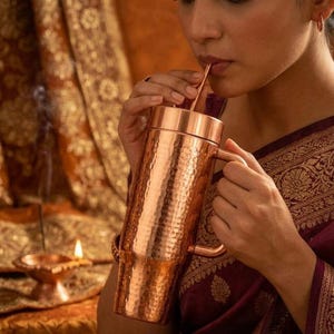 Puede incluir: Un vaso cilíndrico alto de cobre con textura martillada, que incluye una tapa a juego, un asa lateral resistente y una pajita fina de cobre. La superficie metálica refleja la luz, resaltando el acabado con hoyuelos del recipiente.