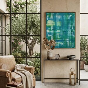 May include: An interior scene featuring a leather armchair with a beige throw blanket, a small wooden stool with a book, and a framed abstract painting with blue and green hues. A black metal console table holds a vase, clock, and bowl. A jute rug and a gold lantern complete the decor.