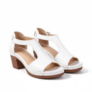 May include: A pair of white T-strap sandals featuring an open-toe design, a secure ankle strap with a metal buckle, and a stacked wooden block heel. The sole includes visible stitching along the edge, providing a structured look to the footwear.