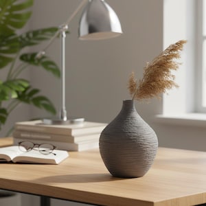 May include: A gray, textured vase with dried ornamental grass sits on a wooden desk. A silver desk lamp and a stack of books are in the background. A pair of glasses rests on an open book.