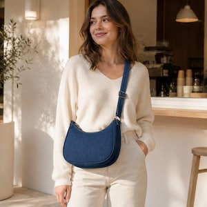 May include: A dark blue denim crescent-shaped crossbody bag with a matching adjustable shoulder strap and a metal buckle. The bag features a curved silhouette, a top zipper closure, and a structured base, worn across the torso with a cream sweater.
