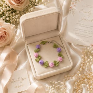 May include: A crochet bracelet featuring alternating light purple and soft pink spherical flower beads connected by small green crocheted leaves. The bracelet includes a silver-toned metal chain with a lobster clasp closure, presented inside a beige velvet box.