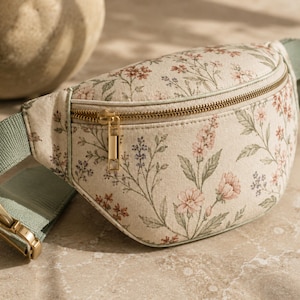 May include: A beige fanny pack featuring a delicate floral pattern in shades of pink, green, and blue. The bag includes a gold-toned metal zipper closure and an adjustable sage green fabric strap with matching gold-toned metal hardware components.