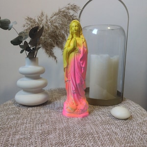 Peut inclure: Une statue vibrante de la Vierge Marie, peinte en jaune fluo et rose, se dresse sur une surface texturée. La statue est ornée d'un chapelet et a les mains jointes en prière. L'arrière-plan comprend un vase blanc avec de la verdure, de l'herbe ornementale séchée et un bougeoir en verre.