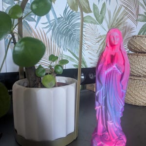 May include: A standing figurine of a person with hands clasped in prayer, featuring a vibrant gradient of neon pink, purple, and blue colors. The figure is depicted in long, flowing robes with a head covering, standing on a small octagonal base.