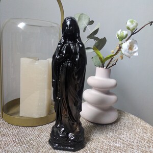 Könnte beinhalten: Eine schwarze Statue einer Figur mit gefalteten Händen, die ein langes, drapiertes Gewand und eine Kopfbedeckung trägt. Die Figur steht auf einem Sockel und hat einen sichtbaren Rosenkranz, der von den Händen herabhängt. Die Statue hat eine glatte Oberfläche.
