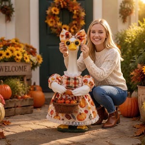 May include: A white goose statue wearing a dress with a pumpkin and autumn leaf pattern, a matching bow, and round sunglasses. The goose holds a small woven basket filled with miniature pumpkins, standing on a stone patio with fall decor.