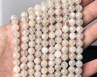 May include: A collection of smooth, round, off-white moonstone beads arranged in multiple vertical strands. Each bead features a subtle, pearlescent sheen and a polished finish, reflecting light across the surface of the spherical gemstone units.