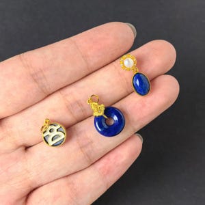 May include: Three gold-toned jewellery charms. One is a circular charm with a white letter B on a dark blue background. The second is a blue lapis lazuli donut-shaped charm with a gold floral bail. The third is an oval blue lapis lazuli pendant.