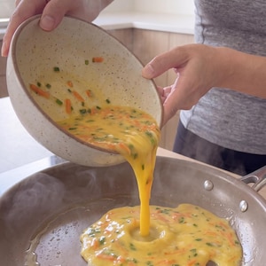 Peut inclure: Une personne verse un mélange d'œufs avec des légumes hachés d'un bol tacheté dans une poêle antiadhésive sur une cuisinière. Le mélange d'œufs est jaune avec des taches vertes et oranges, suggérant une omelette ou des œufs brouillés.