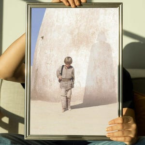 Puede incluir: Fotografía enmarcada de un niño en un paisaje desértico. El niño lleva un atuendo beige y una mochila. La sombra de una figura es visible en la pared detrás de él. El marco es plateado.
