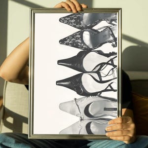 Könnte beinhalten: Schwarz-Weiß-Druck in einem Rahmen mit einer vertikalen Anordnung von High Heels. Die Schuhe variieren im Stil, einschließlich Spitze, Leder und Riemchendesigns. Das Bild wird von einer Person in Jeans gehalten.