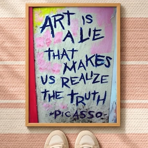 Könnte beinhalten: Ein rechteckiges gerahmtes Wandbild mit hellem Hintergrund und rosa sowie gelben Farbflecken. Der Text in dunkelblauer Tinte lautet: ART IS A LIE THAT MAKES US REALIZE THE TRUTH -PICASSO. Der Rahmen besteht aus hellem, naturfarbenem Holz.