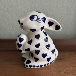 May include: A ceramic rabbit figurine with a glossy cream-colored finish, decorated with a repeating pattern of small, dark blue hearts. The rabbit is depicted in a seated position, holding a matching oval-shaped egg between its front paws.