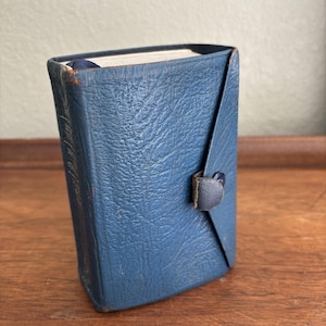 May include: A small, rectangular, blue textured book with a wrap-around cover secured by a single snap button closure. The spine features faint, embossed lettering, and the edges of the cover show signs of wear with visible brown undertones.