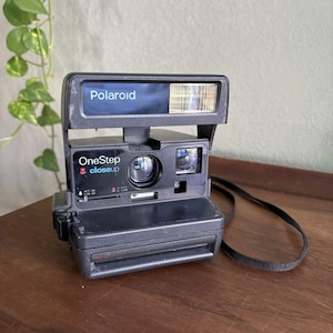May include: A black Polaroid OneStep closeup instant camera featuring a built-in flash, a central lens, and a viewfinder. The front panel displays the text Polaroid, OneStep, and closeup with a small red tulip icon and distance markings.