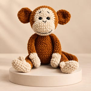 Pode incluir: Um peluche de macaco feito em croché com fio castanho macio e texturizado, com rosto, orelhas, mãos e pés de cor creme. O macaco tem olhos de segurança de plástico preto e uma boca preta bordada, sentado numa posição ereta com uma cauda.