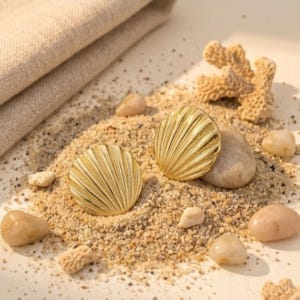 Puede incluir: Dos pendientes dorados en forma de concha marina descansan sobre arena, rodeados de pequeñas piedras y coral. Un paño beige doblado está en el fondo. Los pendientes tienen un diseño acanalado.