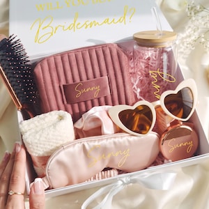 Puede incluir: Una caja de regalo blanca con el texto Will You Be A Bridesmaid? que contiene un neceser malva acanalado, un vaso de cristal con tapa de bambú y pajita, gafas de sol en forma de corazón, un cepillo, una toalla color crema, un antifaz y un espejo.