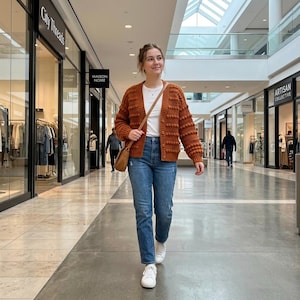 Peut inclure: Un gilet boutonné à manches longues de couleur rouille avec un motif texturé à pompons, porté sur un t-shirt blanc uni avec un jean bleu, des baskets blanches et un sac à bandoulière en cuir marron dans un couloir de centre commercial.