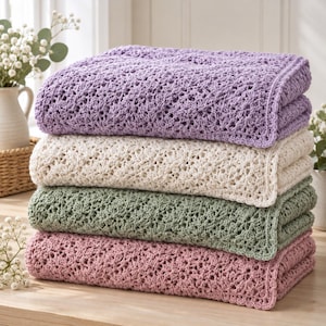 May include: A stack of four folded crochet blankets in pastel colours, including lavender, cream, sage green, and dusty rose. Each blanket features a consistent, intricate openwork floral pattern with a finished edge, neatly layered on a flat surface.