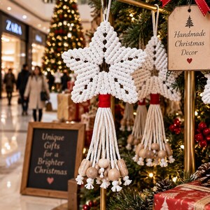 Può includere: Un ornamento a forma di stella in macramè bianco con un avvolgimento di filo rosso sopra una frangia di cordini bianchi decorata con perline di legno naturale. Un'etichetta appesa riporta il testo Handmade Christmas Decor con un'icona di albero e un cuore rosso.