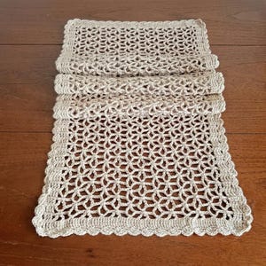 May include: A rectangular beige crochet table runner featuring an intricate openwork star pattern throughout the center. The runner is finished with a solid, dense crochet border that has a scalloped edge along the entire perimeter.