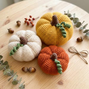 May include: Three crocheted pumpkins in cream, orange, and yellow sit on a wooden surface. Each pumpkin has a brown stem and green vine detail. Acorns, berries, and greenery are scattered around the pumpkins, creating a fall-themed display.