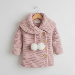 May include: A dusty pink crochet coat featuring a wide collar, long sleeves with folded cuffs, and a front closure with four light brown wooden buttons. Two white pom-poms hang from the collar area on matching crochet strings.