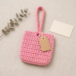 Puede incluir: Una pequeña bolsa cuadrada de ganchillo rosa con un asa trenzada única en la parte superior. Una etiqueta de papel rectangular en blanco de color marrón claro está unida a la parte frontal de la bolsa con un cordel fino de color claro.