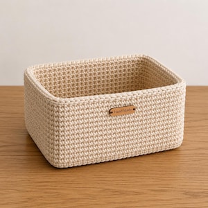 May include: A rectangular storage basket made of cream-colored crocheted cord with a dense, uniform stitch pattern. The basket features a small, horizontal, light brown leather-like tag centered on the front exterior with the text Hand Made.