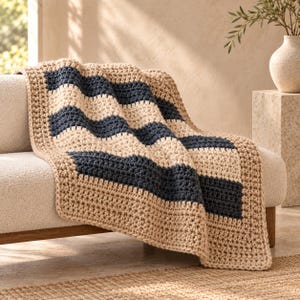 Puede incluir: Una manta de ganchillo gruesa con un patrón repetitivo de anchas rayas horizontales en azul marino oscuro y crema, rematada con un borde beige sólido. La textura del hilo grueso es visible en toda la pieza textil rectangular.