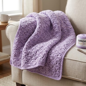 May include: A lavender crochet blanket featuring a repeating shell stitch pattern. Next to the blanket sits a ball of Caron Baby Cakes yarn in the color Lavender and a metal crochet hook. The blanket has a textured surface and soft appearance.