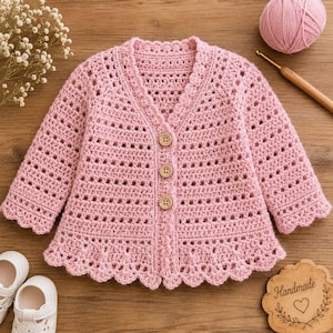 May include: A light pink, handmade crochet cardigan with a scalloped hem and collar. The baby sweater has long sleeves and wooden buttons. A ball of pink yarn and a crochet hook are visible in the background.
