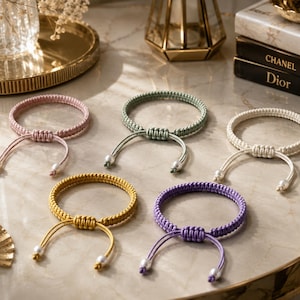 Op de afbeelding: Vijf verstelbare koordarmbanden in roze, saliegroen, crème, mosterdgeel en lavendel. Elke armband heeft een geweven macraméband met een schuifknoopsluiting en twee bungelende koorduiteinden, elk afgewerkt met een kleine witte parelkraal.