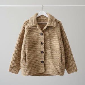 Op de afbeelding: Een beige gehaakt vest met een textuur in wafelsteek, een kraag, lange mouwen en vier donkerbruine knopen aan de voorzijde. Het kledingstuk is voorzien van twee verticale steekzakken op de onderste voorpanden.